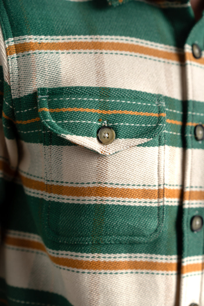 Close-up of a green and beige plaid shirt pocket with a button, highlighting the fabric texture and striped pattern.