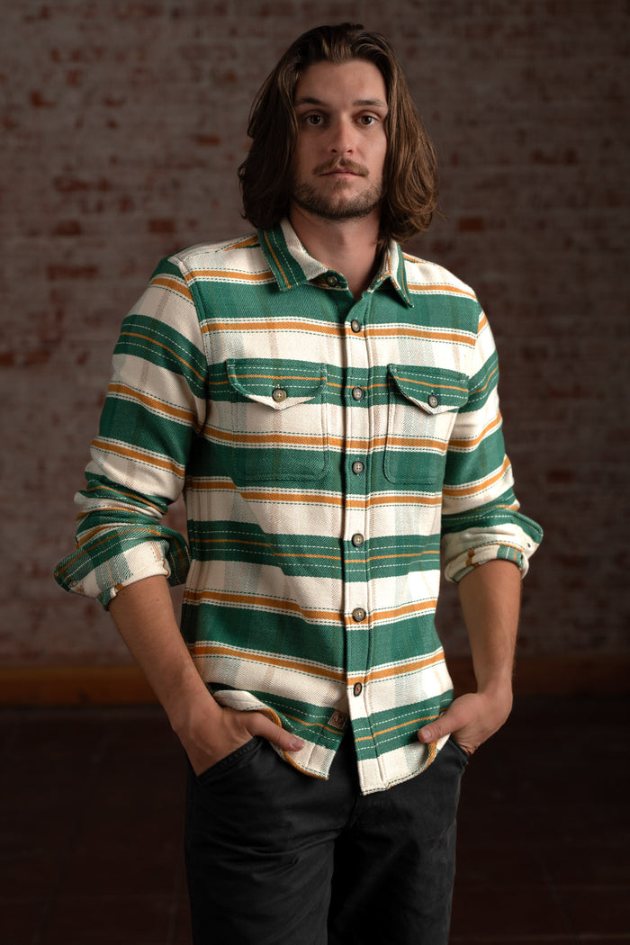 Man in a striped green and white shirt stands confidently against a brick wall, hands in pockets, with a relaxed expression.