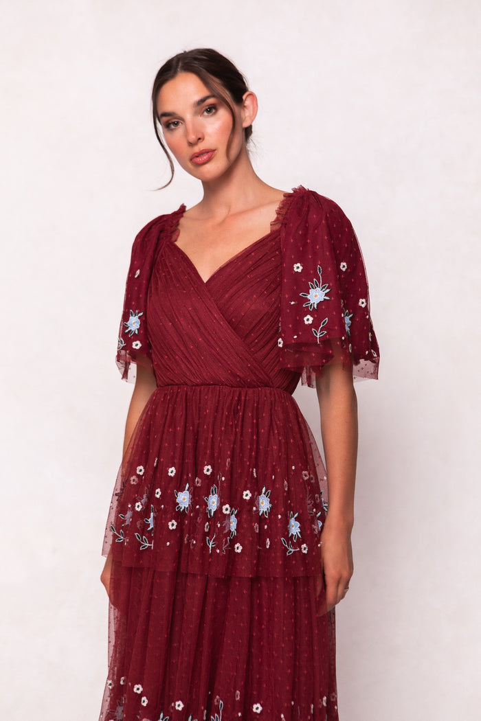 A woman stands against a plain light background wearing the Ivy City Co Mabel Dress in Burgundy, featuring blue and white floral embroidery, sheer puffed short sleeves, and a wrap-style v-neckline.