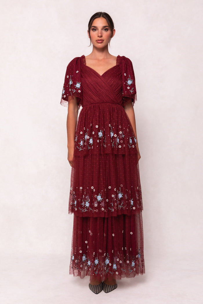 A woman wearing the Mabel Dress in Burgundy by Ivy City Co—a burgundy maxi dress featuring short sleeves and blue floral embroidery—stands against a plain light background.