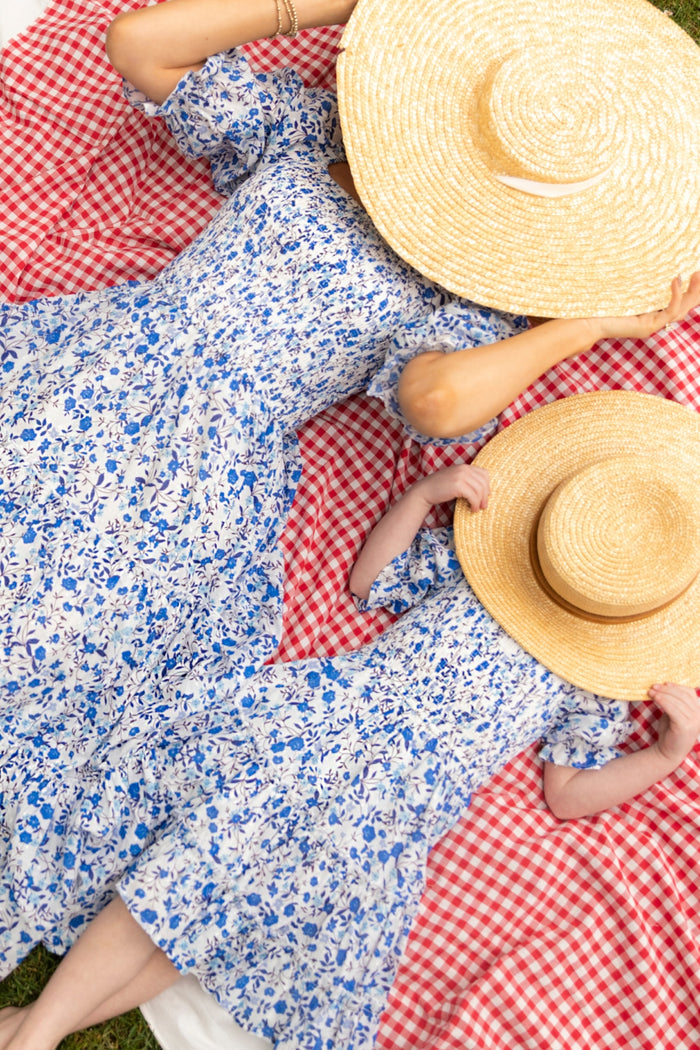 A woman and child in matching Ivy City Co Madeline Dress in Ditsy Cobalt Floral lounge on a red and white blanket, straw hats over their faces. These cotton midi dresses are perfect for maternity, nursing, and sunny days outdoors.