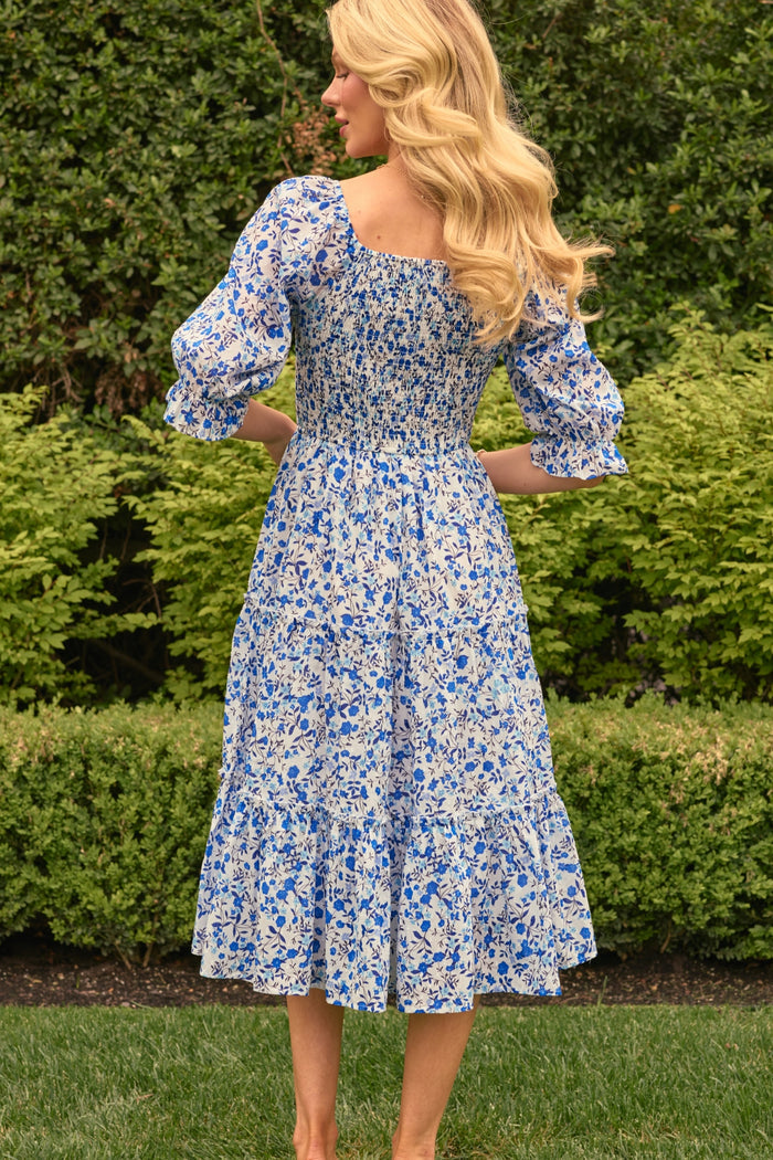 A woman with long blonde hair stands on grass, facing away, wearing the Ivy City Co Madeline Dress in Ditsy Cobalt Floral—a blue and white cotton midi that’s maternity and nursing friendly—amid green bushes and trees.
