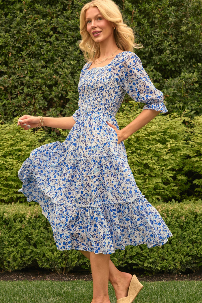 A woman with blonde hair stands on grass, smiling and holding her skirt while wearing the Ivy City Co Madeline Dress in Ditsy Cobalt Floral. Lush green hedges and bushes are visible in the background.