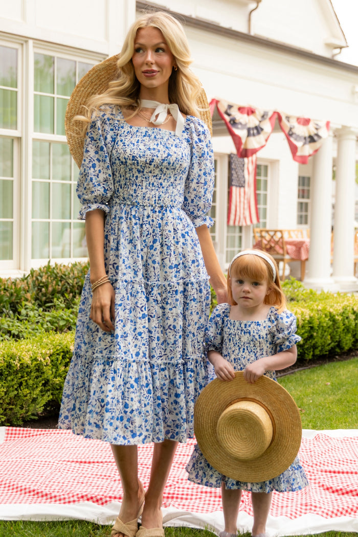 A woman and girl wear matching Ivy City Co Madeline Dresses in Ditsy Cobalt Floral, standing on a picnic blanket outdoors. Each holds a straw hat, with patriotic bunting decorating the house behind them.