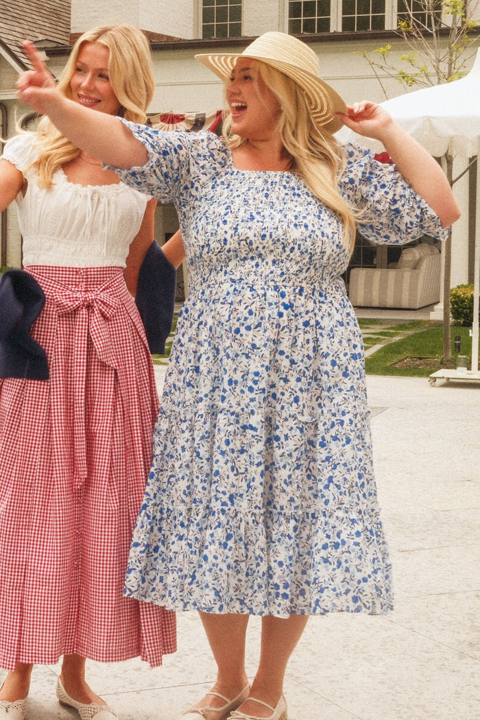 Two women smile outdoors. One wears the Ivy City Co Madeline Dress in Ditsy Cobalt Floral with a straw hat, while the other wears a white top with a red plaid skirt and bow. A house and tent appear in the background.