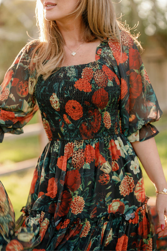 A woman stands outdoors in sunlight, wearing the Ivy City Co Madeline Dress in Imogen Moody Floral—a sheer, flowy black gown featuring a smocked bodice and a vibrant red, orange, and green floral print. Her hair falls loose around her shoulders, complemented by a delicate heart-shaped necklace. This elegant style from Ivy City Co is also maternity friendly.
