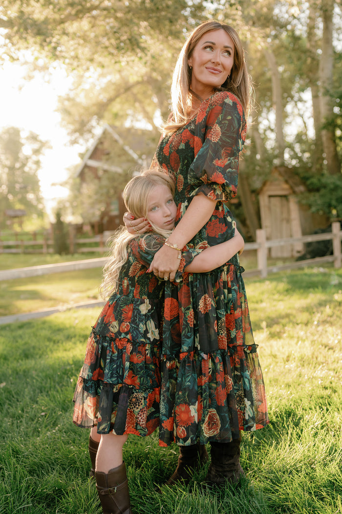 A woman and a young girl, both wearing the Ivy City Co Madeline Dress in Imogen Moody Floral with smocked bodices, stand together on grass in a sunlit yard. The girl hugs the woman’s waist as they share a peaceful moment. Trees and a rustic building in the background highlight the maternity-friendly elegance of their coordinated outfits.