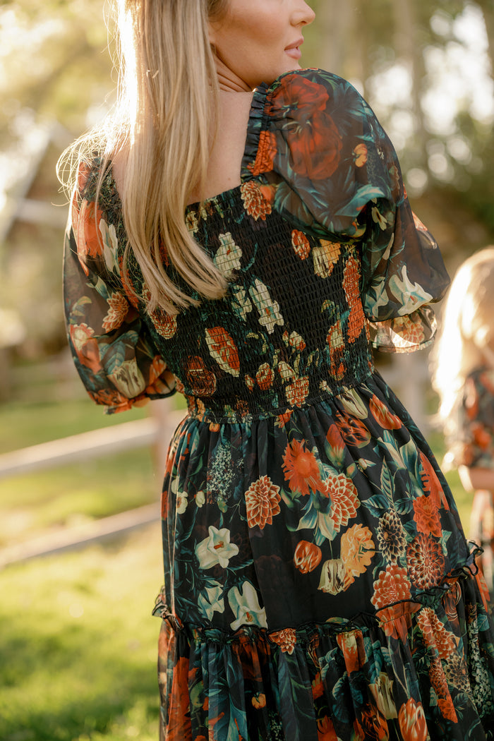 A woman with long blonde hair wears the Ivy City Co Madeline Dress in Imogen Moody Floral, featuring a smocked bodice and vibrant floral patterns. Maternity friendly, the dress combines style and comfort as she stands outdoors in sunlight against a blurred backdrop of grass, trees, and a wooden fence.