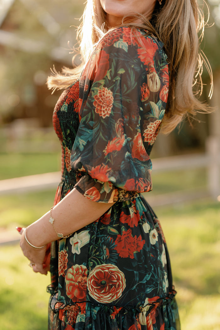 A woman stands outdoors wearing the Ivy City Co Madeline Dress in Imogen Moody Floral, a sheer design featuring red, orange, and green patterns. The smocked bodice provides a flattering, maternity-friendly fit as sunlight highlights her hair while she holds something in her hand. The softly blurred background includes greenery and a wooden fence.