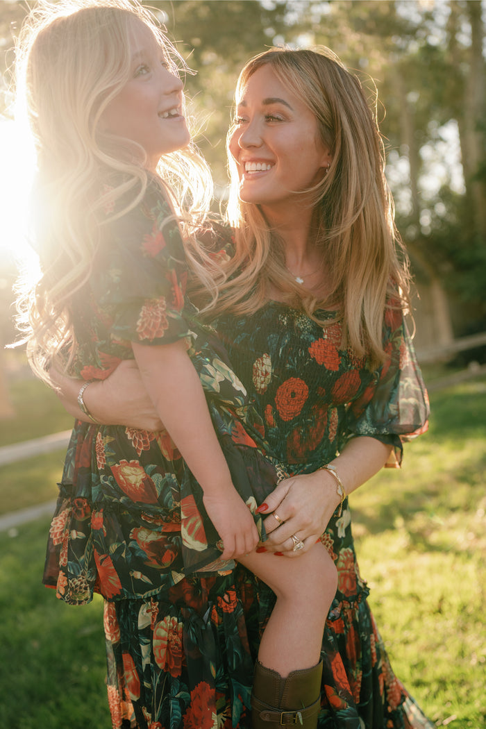 A woman smiles while holding a young girl; both wear matching Ivy City Co Madeline Dresses in Imogen Moody Floral, featuring smocked bodices, as they stand in sunlight on a grassy lawn. These maternity-friendly dresses are perfect for mothers and daughters to enjoy together.