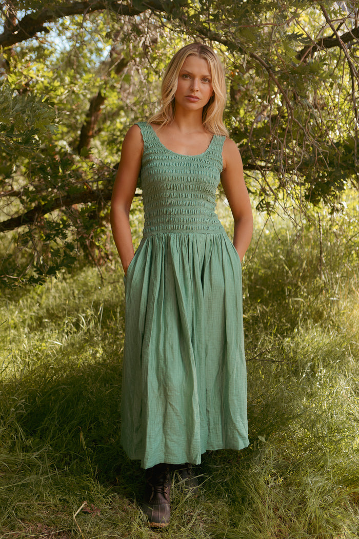 A woman stands outdoors among green trees and grass, wearing the Ivy City Co Marlow Dress in Green—a sleeveless, maternity-friendly midi with a smocked bodice and pockets. Sunlight filters through the leaves, creating a serene atmosphere.