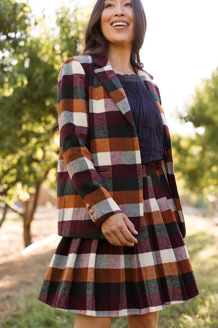 A woman is outdoors, smiling and wearing the Ivy City Co Marren Blazer in Maize Brushed Gingham paired with a matching pleated skirt in autumn hues, layered over a dark top. Trees can be seen softly blurred in the background.