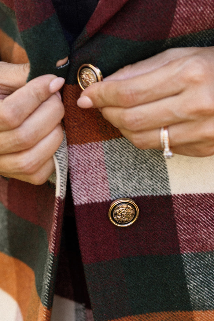 Close-up of hands buttoning the Ivy City Co Marren Blazer in Maize Brushed Gingham, a standout women’s blazer featuring green, burgundy, white, and orange sections in a checkered pattern with gold buttons. One hand wears a ring with a clear stone.