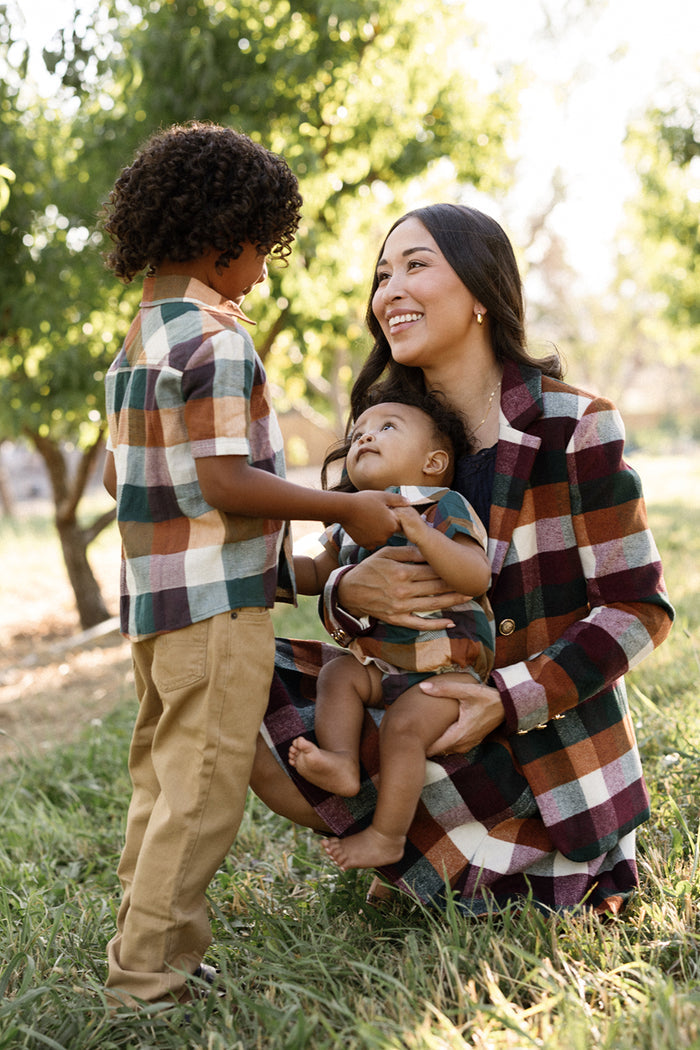 A smiling woman kneels on the grass, holding a baby, while a young boy stands nearby. All three wear matching plaid outfits, and the woman completes her look with the Ivy City Co Marren Blazer in Maize Brushed Gingham. Trees and sunlight are in the background.