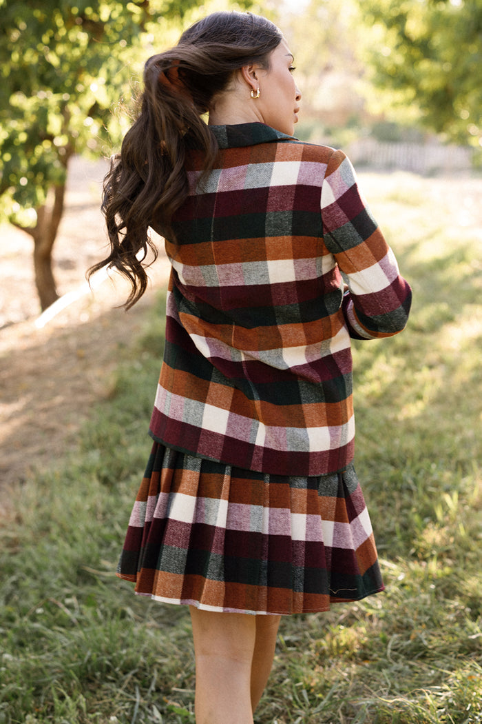A woman with long brown hair walks outdoors, wearing the Ivy City Co Marren Blazer in Maize Brushed Gingham. She is facing away, surrounded by green grass and trees.
