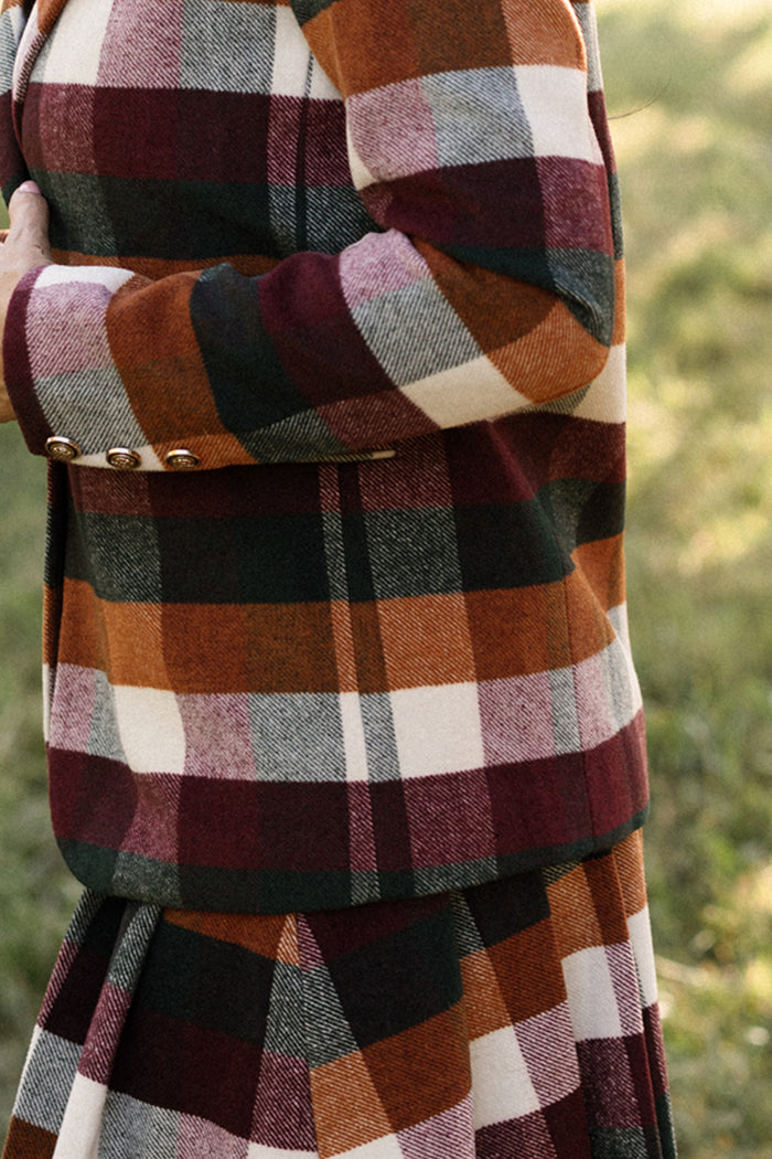 A person wearing the Ivy City Co Marren Blazer in Maize Brushed Gingham, styled with a matching skirt in shades of burgundy, orange, white, and black, stands outdoors on grass, shown from the side.