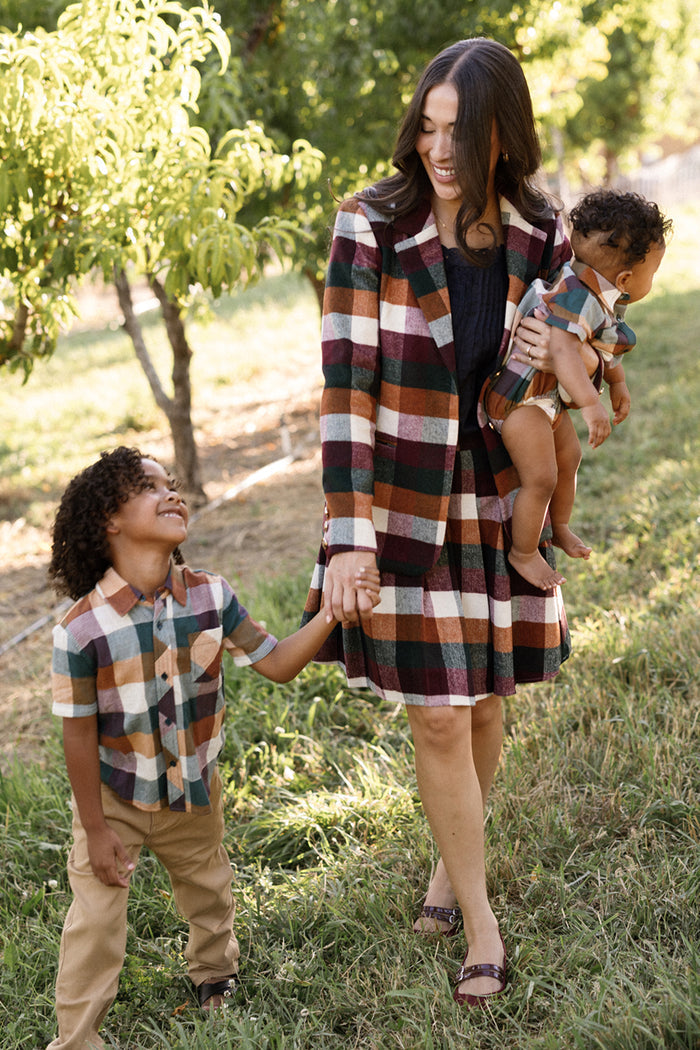 Wearing the Ivy City Co Marren Blazer in Maize Brushed Gingham, a woman holds a baby in one arm and the hand of a smiling young boy as they walk together along a grassy, tree-lined path.
