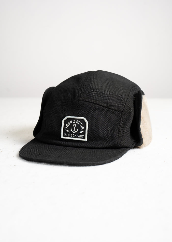 Black cap with ear flaps, featuring "Iron & Resin MFG Company" logo on front, displayed on a white surface.
