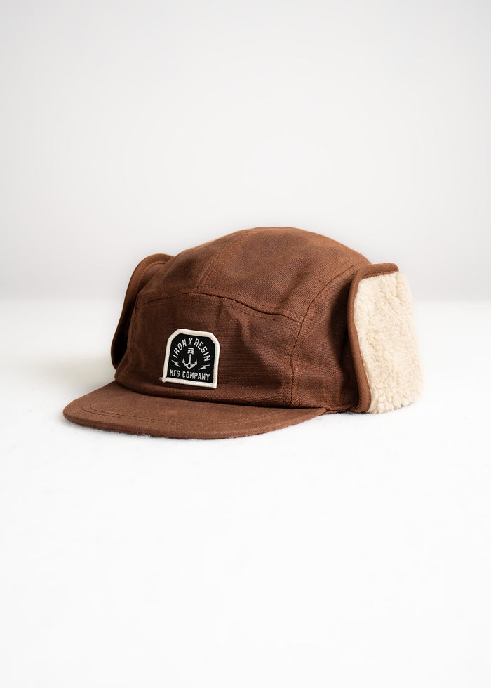 Brown winter cap with ear flaps, featuring a black and white "Iron and Resin MFG Company" patch on the front, on a white background.