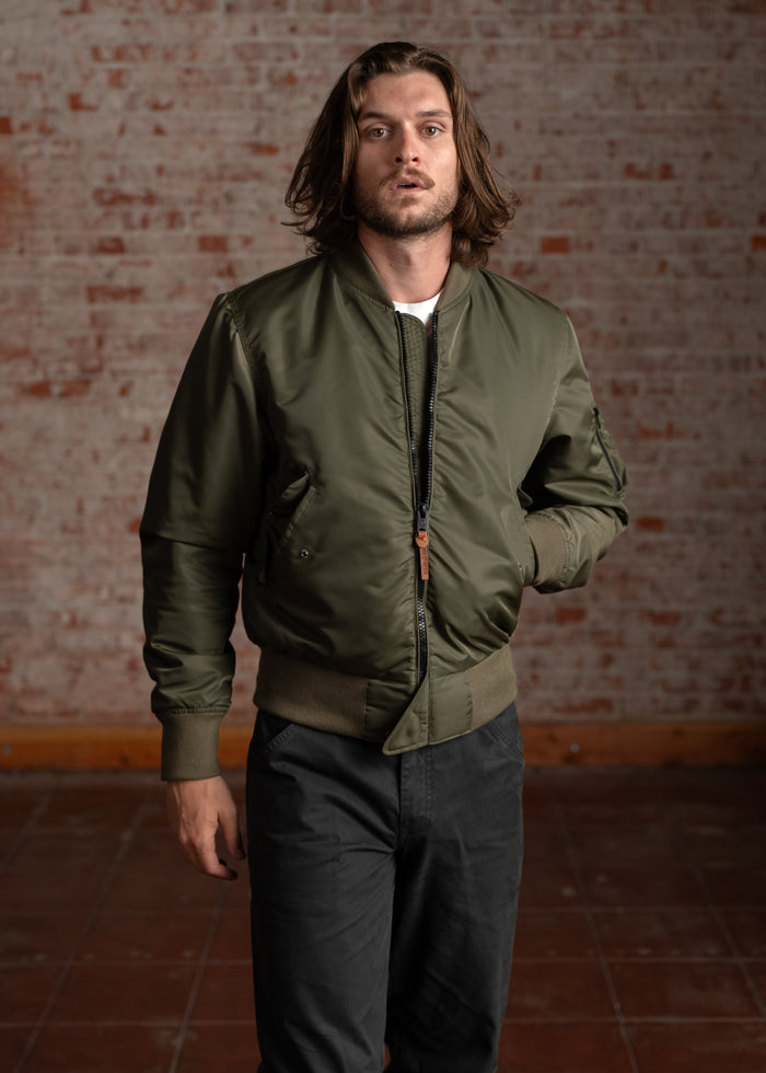 Man in a green bomber jacket stands against a brick wall, looking forward. Casual style with long hair and relaxed posture.
