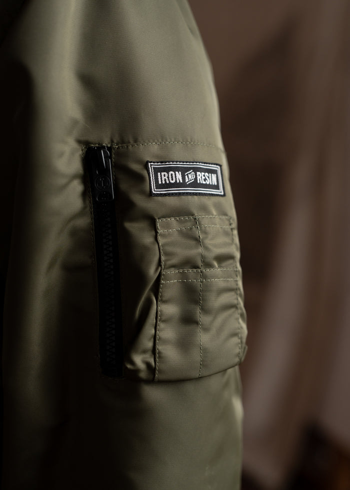 Close-up of an olive green jacket sleeve with a black zipper and "Iron and Resin" patch, highlighting its rugged style.