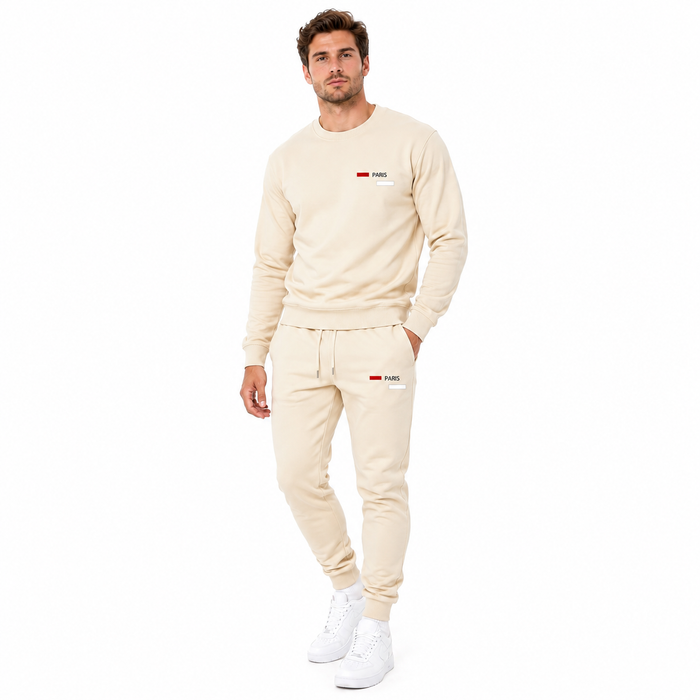 A man modeling a matching beige sweatshirt and jogger pants set with small red and white logo details on the chest and thigh, paired with white sneakers, against a plain white background.