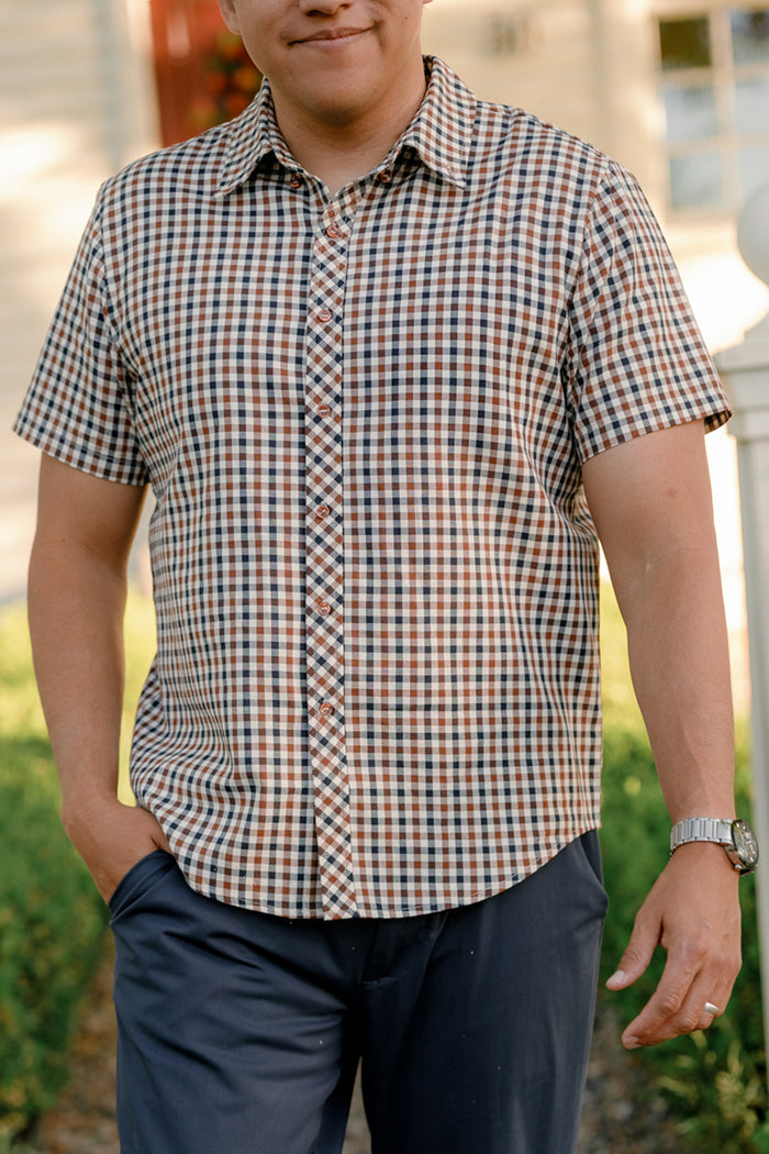 A man stands outdoors with one hand in his pocket and a watch on his wrist, wearing the Ivy City Co Mens James Shirt in Chestnut Gingham—an ideal short-sleeve option for fall layering—paired with dark pants.