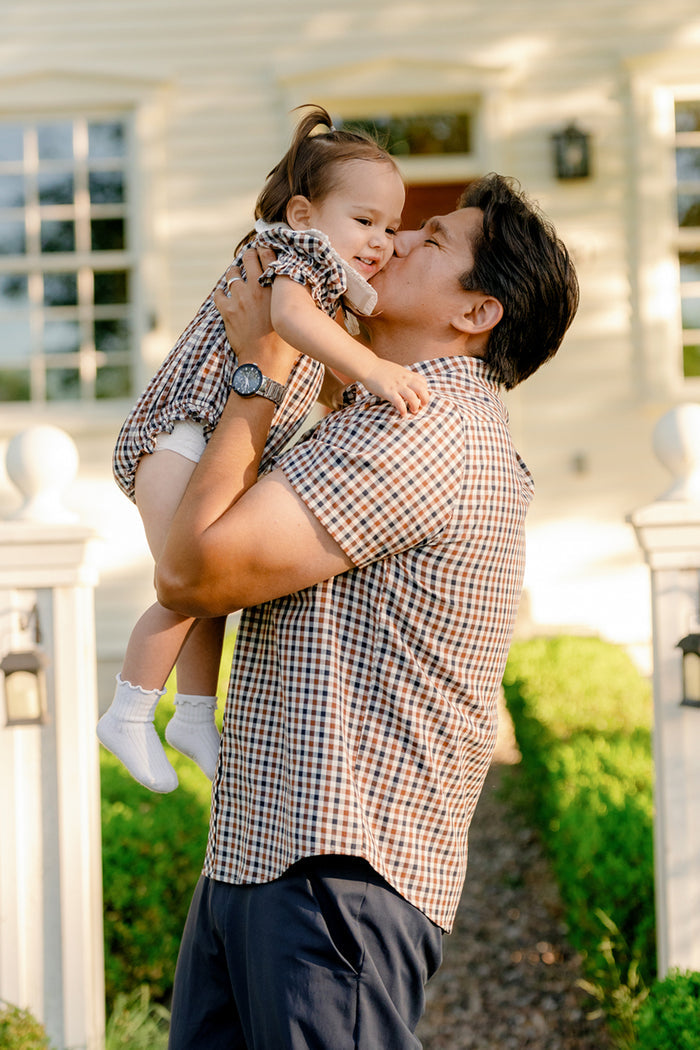 A man lifts a smiling toddler and kisses their cheek outside. Both wear matching plaid outfits perfect for fall layering, with the man in the Ivy City Co Mens James Shirt in Chestnut Gingham. The background shows a bright house and greenery.