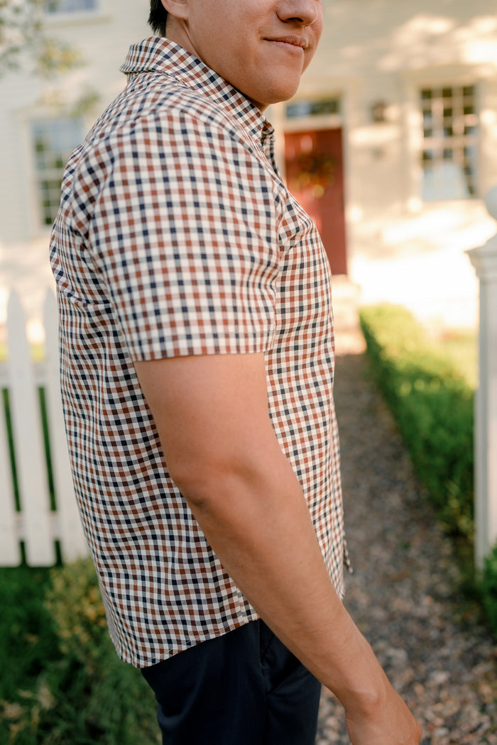 A man wearing the Ivy City Co Mens James Shirt in Chestnut Gingham stands outdoors on a gravel path near a white picket fence and a house with a red door, smiling slightly. This classic cotton shirt is perfect for effortless fall layering.