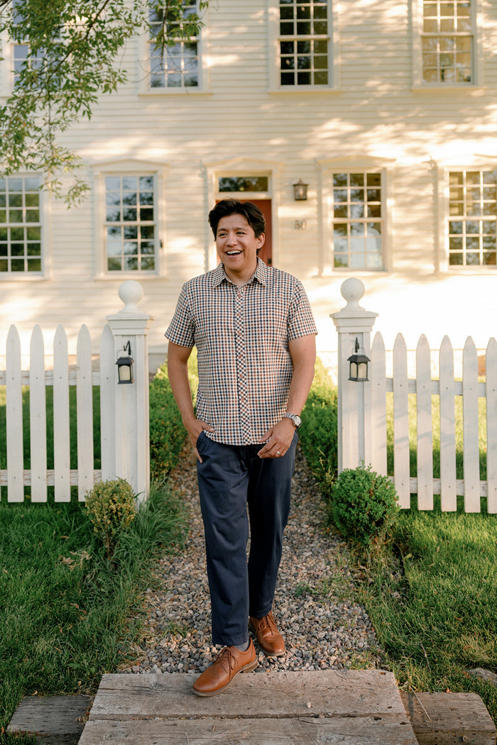Wearing the Ivy City Co Mens James Shirt in Chestnut Gingham and blue pants, a man smiles as he strolls along a stone path outside a white house with a picket fence and lush green lawn, effortlessly showcasing fall layering with his classic cotton shirt.
