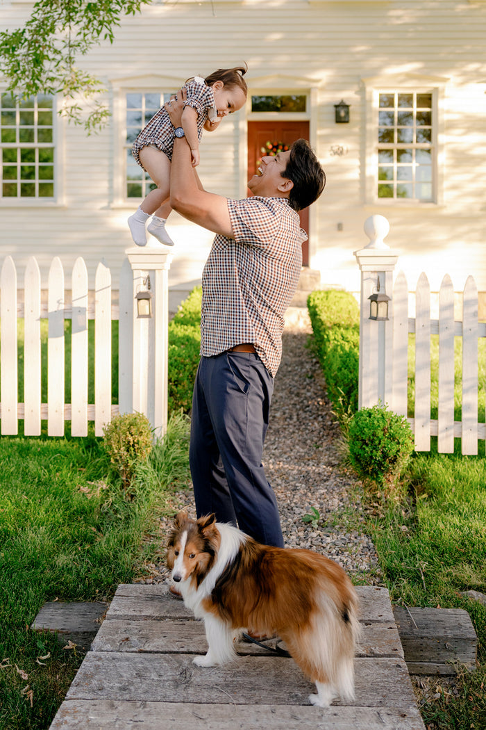 A man wearing the Ivy City Co Mens James Shirt in Chestnut Gingham lifts a smiling child dressed in coordinating outfits outside a white house with a picket fence, both styled perfectly for fall layering. A brown and white dog stands on a wooden path in the foreground.