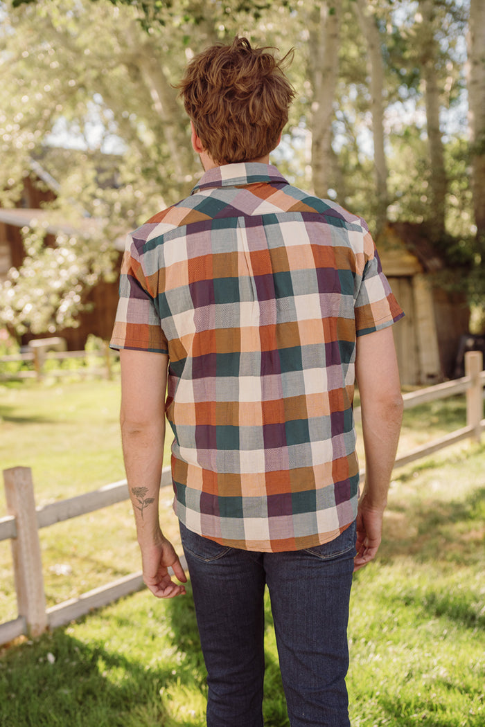 A person stands outdoors facing away, wearing the Ivy City Co Mens James Shirt in Maize Gingham—a short-sleeved, collared cotton-blend shirt—paired with jeans. The background features green grass, trees, and a wooden fence in bright sunlight.