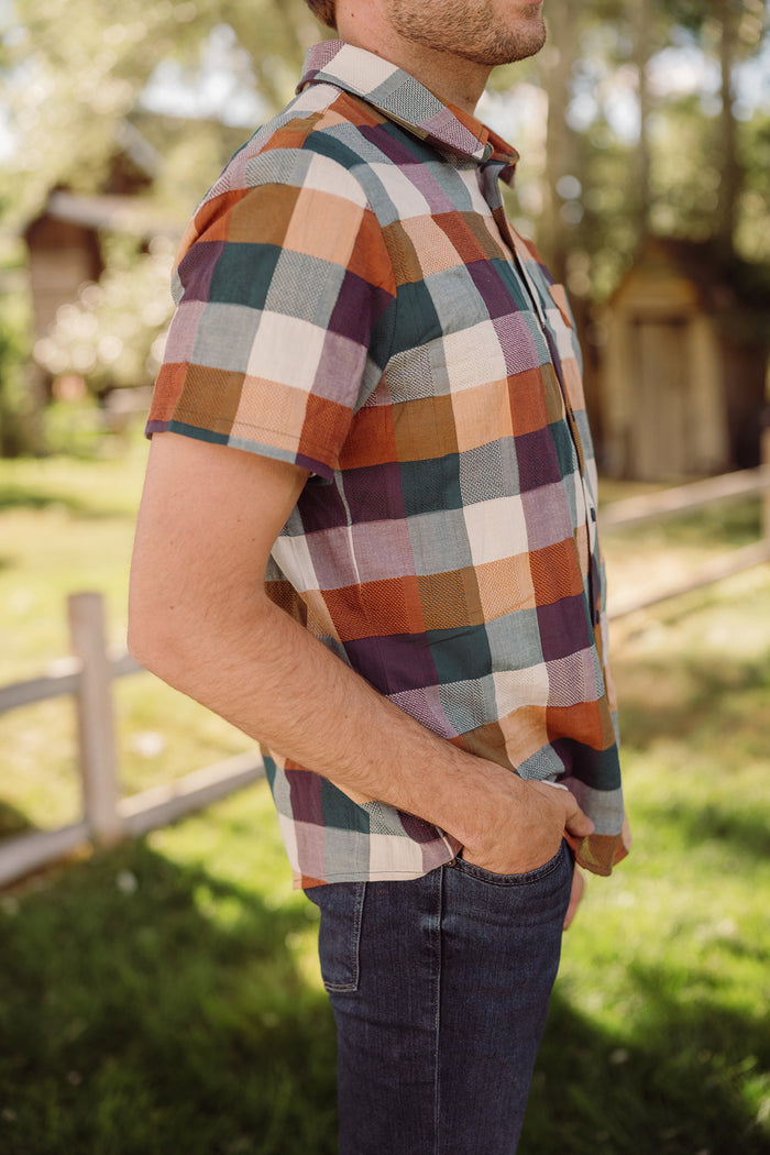 A person stands outdoors with hands in jean pockets, wearing the Ivy City Co Mens James Shirt in Maize Gingham—a short-sleeve, cotton-blend shirt featuring a classic collar neckline. The background features green grass, trees, and a wooden fence.