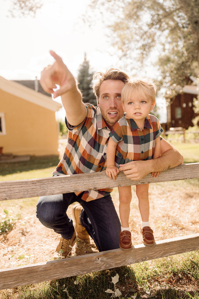 A man kneels by a wooden fence, pointing into the distance, while a young child stands beside him. Both are wearing matching Ivy City Co Mens James Shirts in Maize Gingham with classic collar necklines, adding a timeless touch to their look. The scene is outdoors on a sunny day with houses and trees in the background.