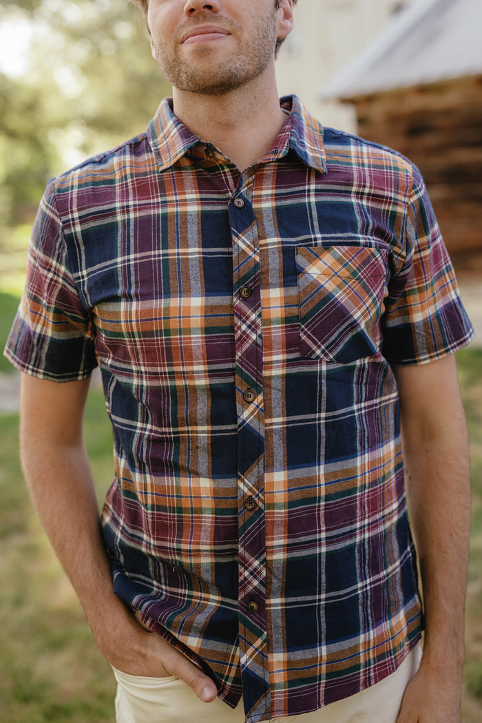 A person is seen outdoors, wearing the Ivy City Co Mens James Shirt in Midnight Plaid—a short-sleeve, multicolored cotton plaid shirt. With one hand in their pocket and a blurred backdrop of greenery and a wooden building, this stylish look is an ideal choice for refreshing your fall wardrobe.