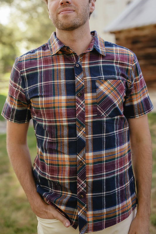 A person is seen outdoors, wearing the Ivy City Co Mens James Shirt in Midnight Plaid—a short-sleeve, multicolored cotton plaid shirt. With one hand in their pocket and a blurred backdrop of greenery and a wooden building, this stylish look is an ideal choice for refreshing your fall wardrobe.