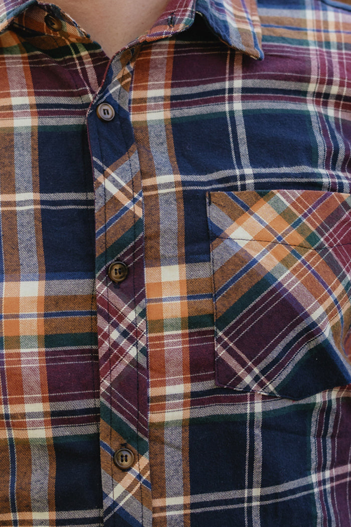 A close-up of a person wearing the Ivy City Co Mens James Shirt in Midnight Plaid, a buttoned cotton shirt with a chest pocket on the right side in shades of navy blue, orange, brown, and white—perfect for elevating your fall wardrobe.