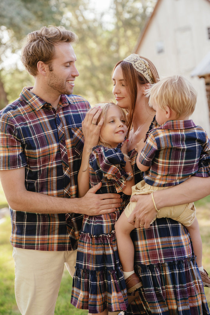 A smiling family of four stands outdoors, wearing matching plaid outfits ideal for any fall wardrobe. The mother holds a young boy, while the father embraces a girl and is dressed in the Ivy City Co Mens James Shirt in Midnight Plaid, crafted from soft cotton. All look happy and close together in a sunlit setting.