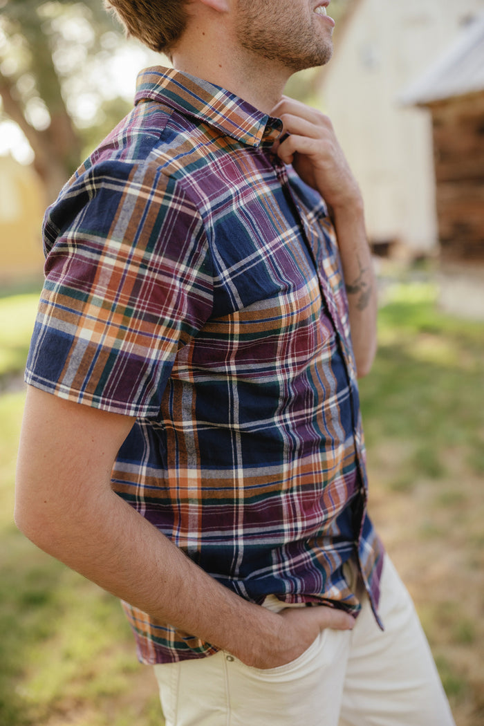 A side profile of a man with light stubble wearing Ivy City Co’s Mens James Shirt in Midnight Plaid—a short-sleeved cotton shirt featuring navy, burgundy, and gold plaid—paired with cream-colored pants. He stands outdoors with one hand tucked into his pocket and the other near his collar, showcasing this timeless piece that’s perfect for any fall wardrobe.
