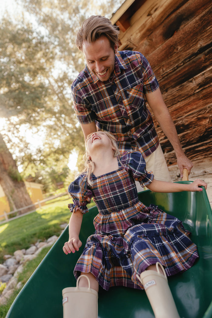 A smiling man, dressed in the Ivy City Co Mens James Shirt in Midnight Plaid—an essential for your fall wardrobe—pushes a laughing young girl down a slide; both wear matching plaid outfits, and the girl sports beige rain boots. They are outdoors near a wooden building, with sunlight streaming through the trees.