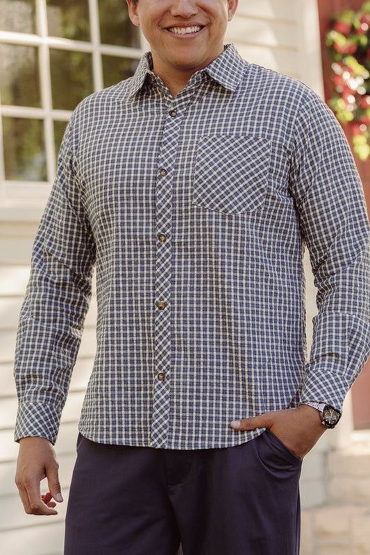 A man stands outside in front of a white building, smiling, with one hand in his pocket. He is wearing the Mens John Shirt in Prussian Blue Plaid by Ivy City Co—a blue and white checkered cotton shirt with a collar neckline—and dark pants.