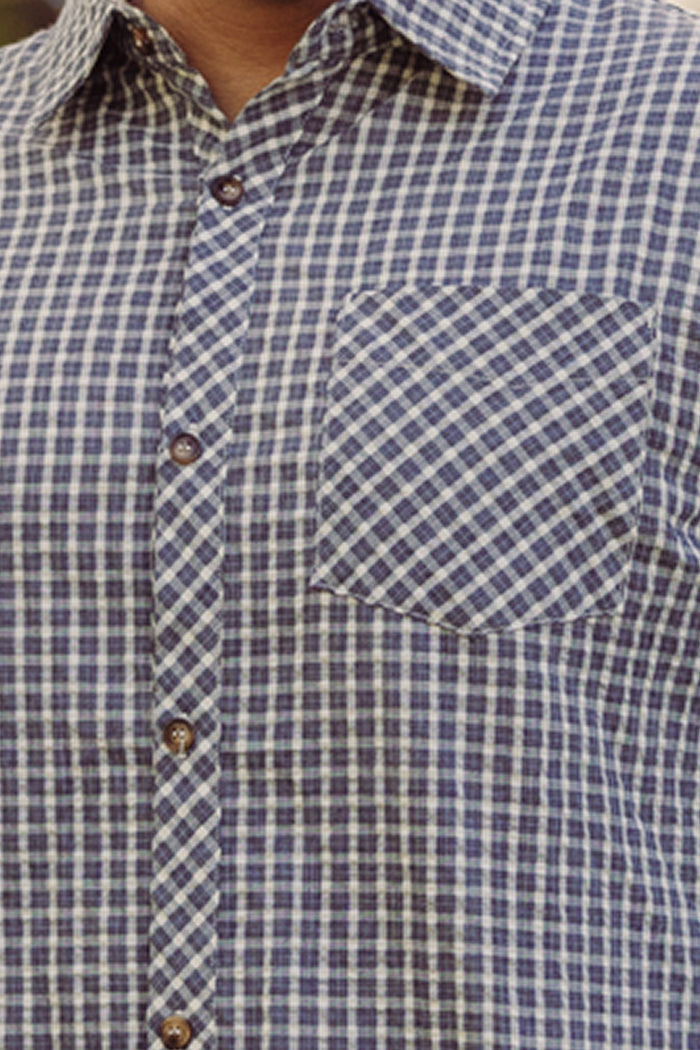 A close-up of a person wearing the Ivy City Co Mens John Shirt in Prussian Blue Plaid, featuring a classic collar neckline and a front pocket on the left side.