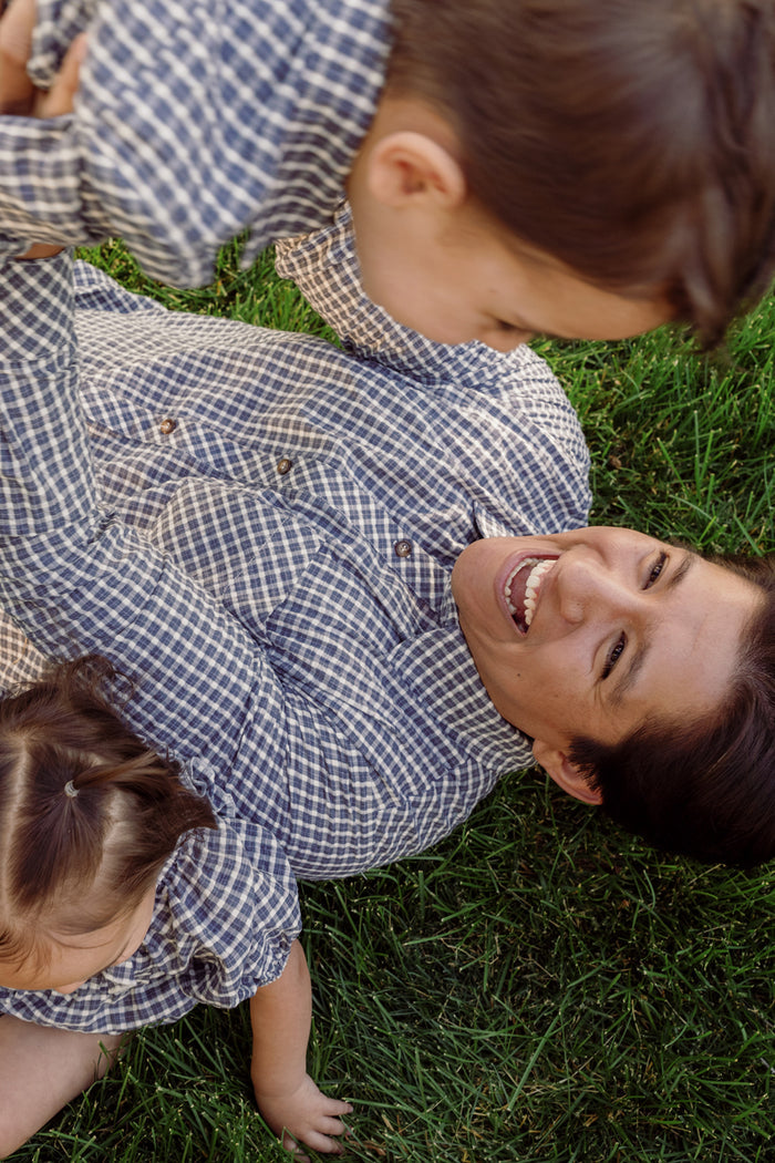 A woman lies on the grass, smiling up at two young children as they share a joyful moment outdoors. All three wear matching Ivy City Co Mens John Shirts in Prussian Blue Plaid, featuring classic collar necklines.