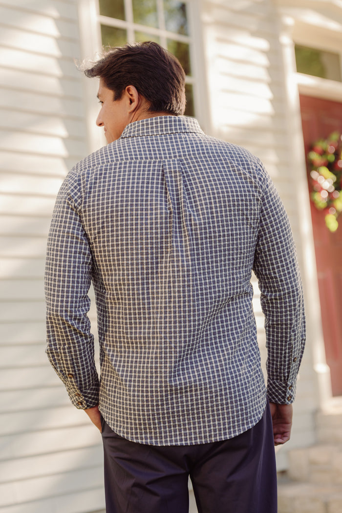 A person wearing the Ivy City Co Mens John Shirt in Prussian Blue Plaid—a checked, long-sleeve cotton shirt with a collar neckline—and dark pants stands outside, facing away from the camera, near a light-colored building where sunlight and shadows play across the wall.