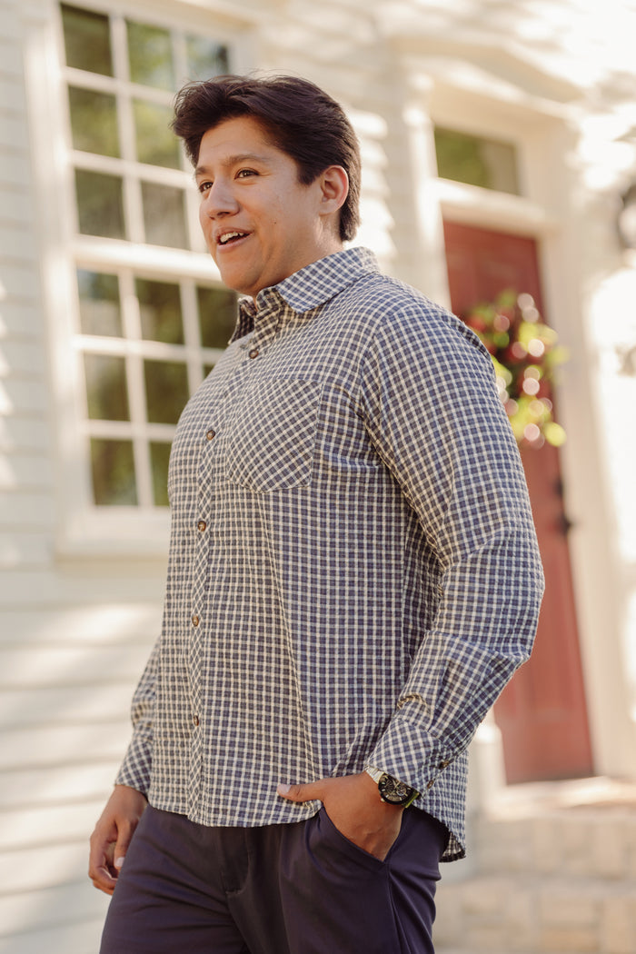 A man wearing the Mens John Shirt in Prussian Blue Plaid by Ivy City Co, featuring a classic collar and blue-and-white checkered cotton fabric, stands smiling outside in front of a house with white siding, a red door, and large windows. He is looking to the side.
