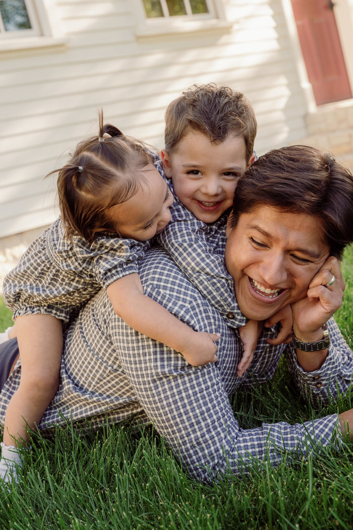 A man in the Ivy City Co Mens John Shirt in Prussian Blue Plaid lies on the grass, smiling, while a young boy and a toddler—each wearing matching plaid outfits with collar necklines—playfully climb on his back in front of a house.