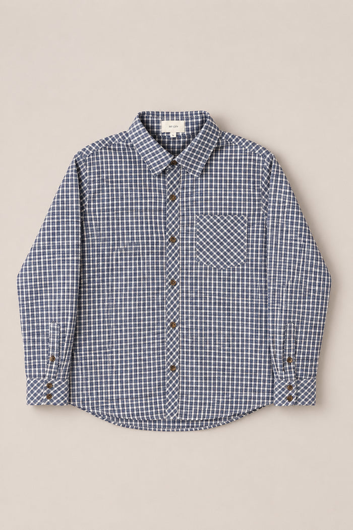The Mens John Shirt in Prussian Blue Plaid from Ivy City Co is crafted from soft cotton and features a collar neckline, long sleeves, a front chest pocket, and brown buttons. Presented laid flat on a neutral background for a classic look.