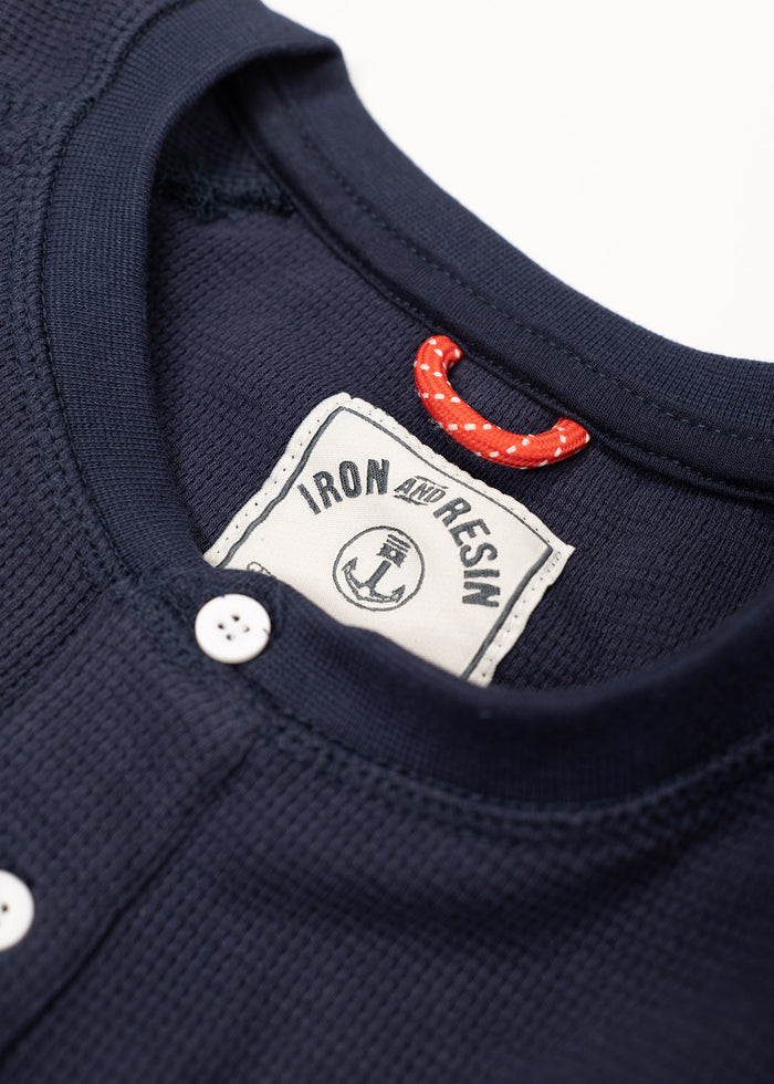 Close-up of a navy waffle-knit shirt with white buttons, featuring an Iron and Resin label and a red polka dot loop inside the collar.