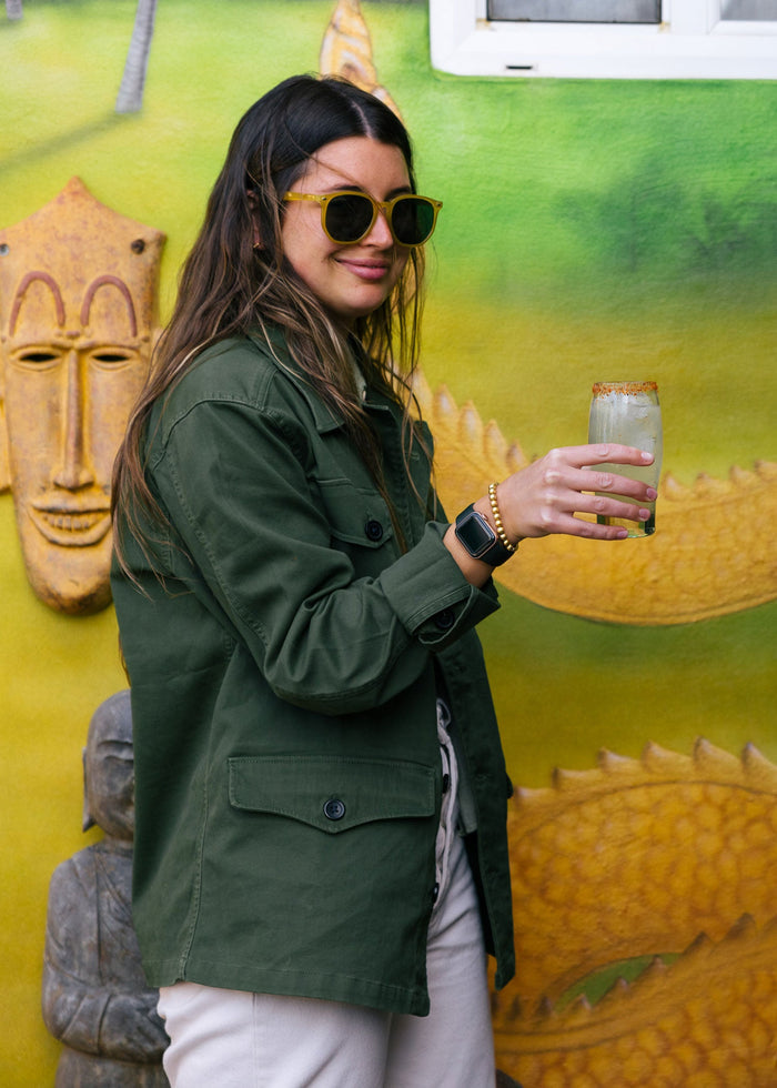 Woman in green jacket and sunglasses holds a drink, standing by a colorful mural with tribal mask and statue.