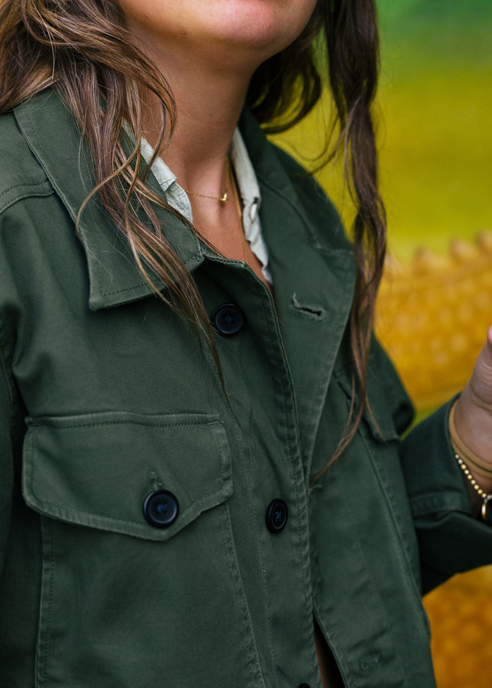 Woman in green jacket with black buttons, holding a drink. Background is blurred with yellow and green hues. Casual setting.